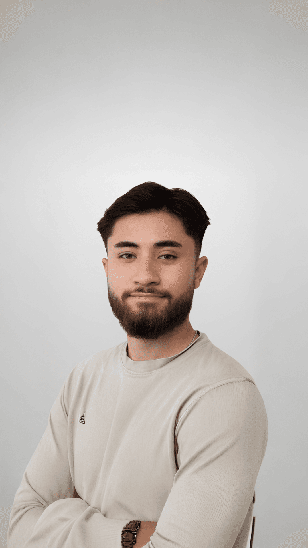 Junger Mann mit Bart und dunklen Haaren im beigen Shirt vor einem schlichten grauen Hintergrund.